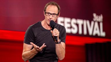 ince Ebert steht in der Mitte des Bildes. Er hebt seine rechte Hand. In der linken Hand hält er sein Mikrofon. Der Hintergrund ist rot beleuchtet.