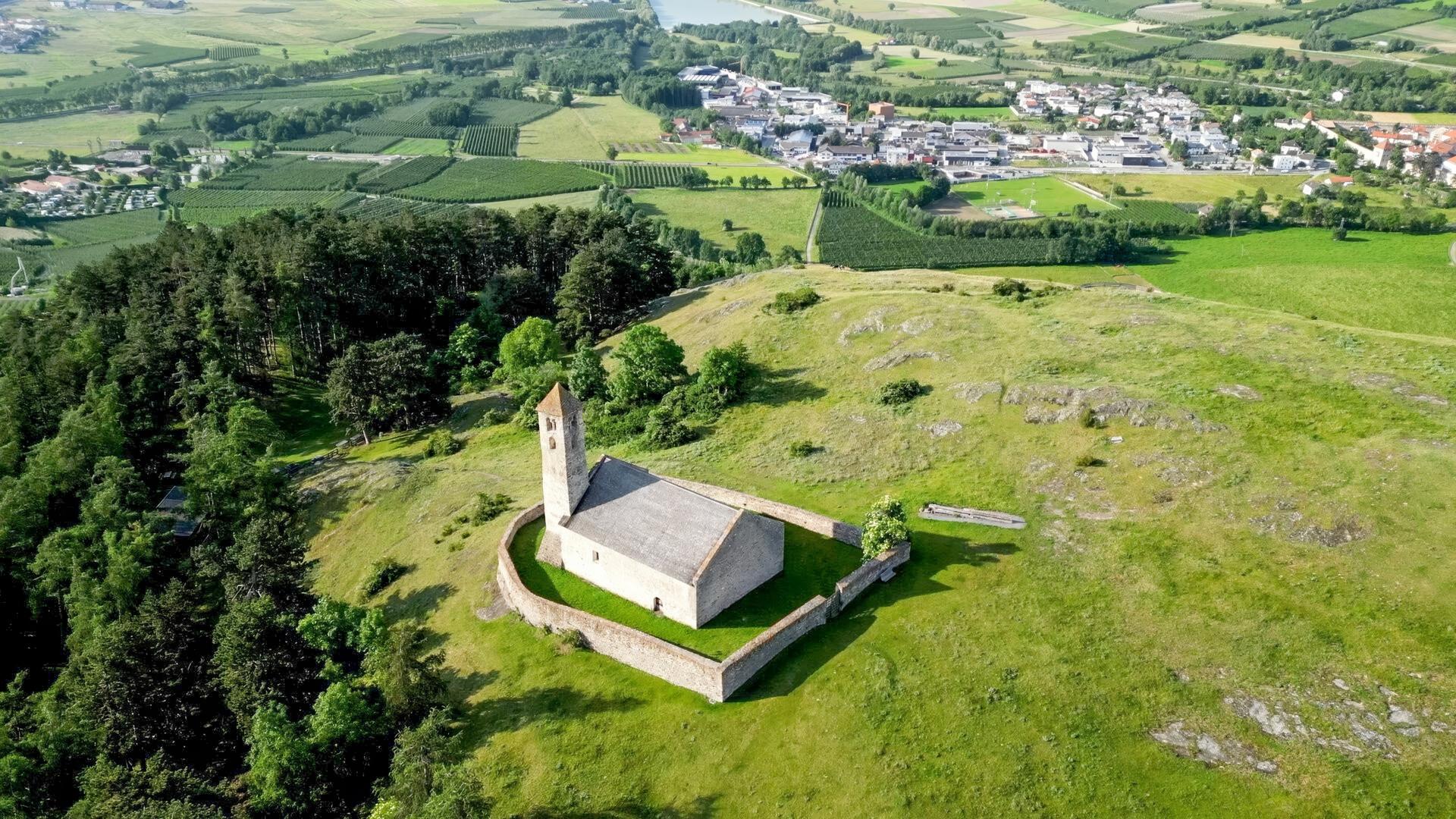 Das Bild zeigt die kleine Kirche Tartscher Buehel, die auf einem sanften Hügel steht. Sie hat eine rechteckige Form mit einem schlichten grauen Dach und einem Glockenturm, der an einer Ecke erhöht ist. Um die Kirche herum befindet sich eine halbkreisförmige Steinmauer. Der Hügel ist von saftigem Grün umgeben, und es gibt einige verstreute Bäume. Im Hintergrund erstreckt sich eine weite Landschaft mit Feldern und Wäldern, die in verschiedenen Grüntönen leuchten. In der Ferne sind einige Gebäude und Straßen zu erkennen, die Teil einer Ortschaft sind. Das Bild vermittelt einen ruhigen, natürlichen Eindruck und zeigt die idyllische alpine Umgebung des Vinschgaus in Südtirol.