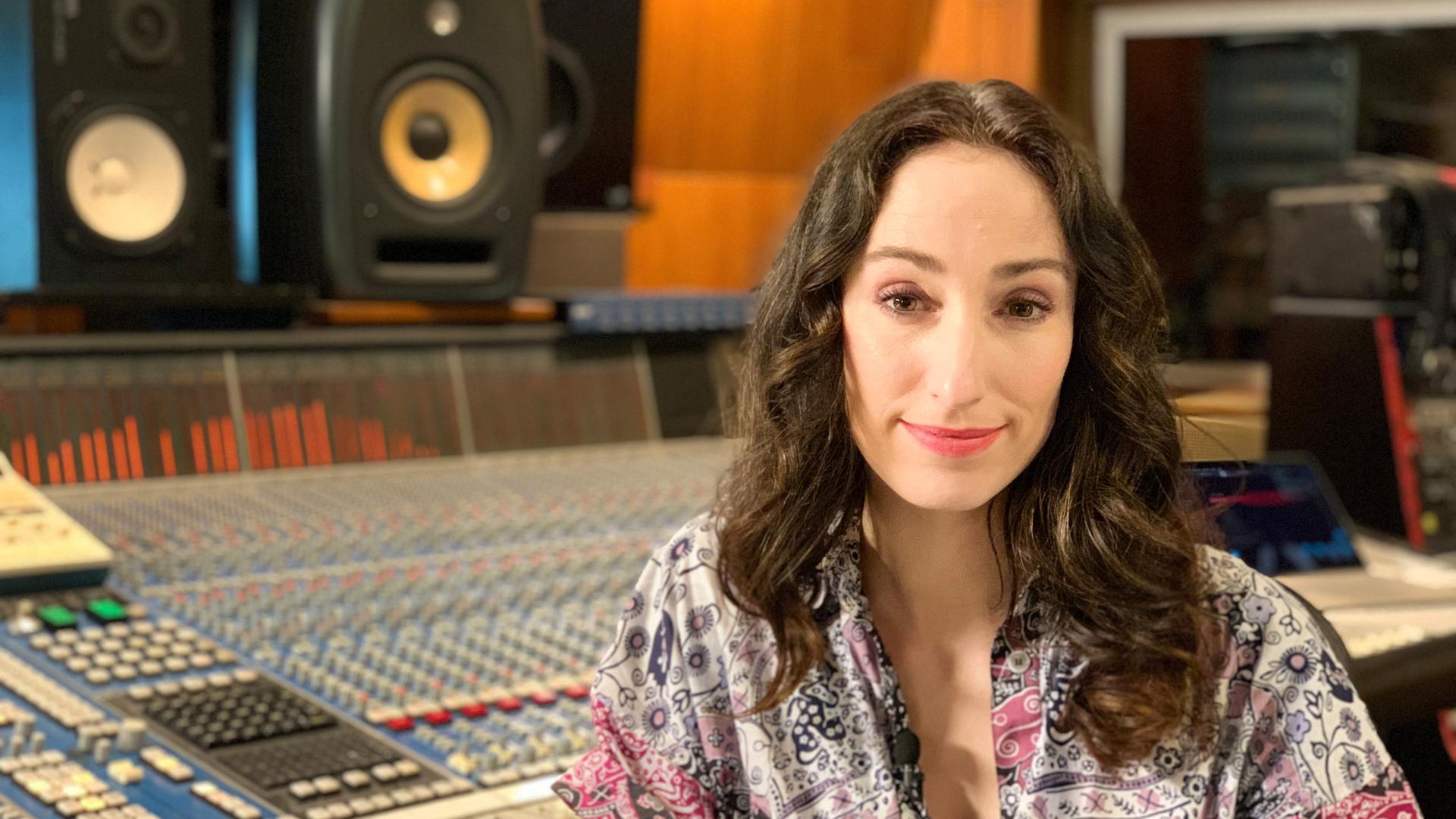 Eine Frau mit langen dunklen Locken sitzt in einem gemusterten Kleid vor einem großen Mischpult, im Hintergrund stehen zwei Boxen. 