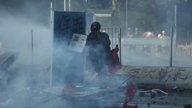 "Voices from Hong Kong": Straßenszene von einer Demonstration in Hongkong: Ein Wasserwerfer, der nicht im Bild zu sehen ist, wird gegen Demonstranten eingesetzt. Ein Mann, der dunkle Kleidung und einen Helm trägt, sucht hinter einem Schild Schutz. Er hat ein kleines Schild in der Hand, auf dem „End“ steht. Auf der Straße liegen Absperrgitter und Metallgegenstände verstreut herum.