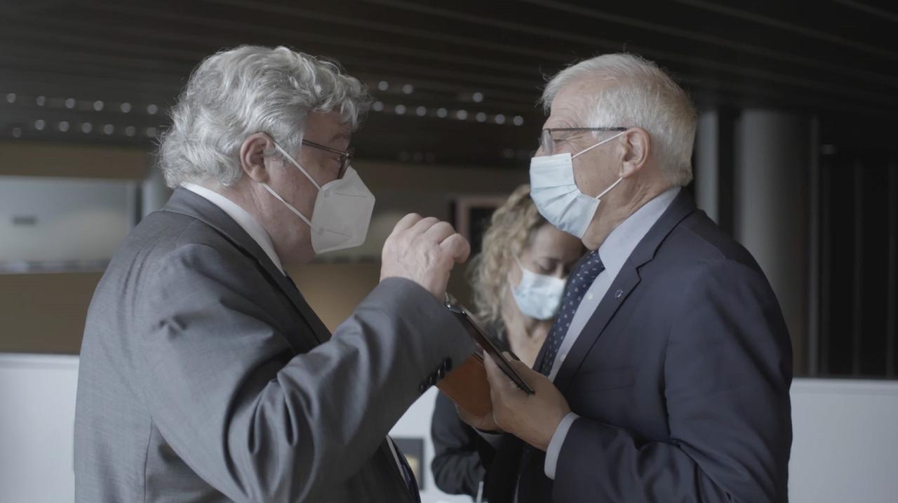 Zwei ältere, weißhaarige Männer in Anzug und Schlips, die beide FFP 2-Masken tragen, sind auf dem Gang eines Gebäudes in ein lebhaftes Gespräch vertieft.