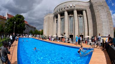 Menschen schwimmen in und stehen an einem Schwimmbad vor einem Theaterbau.