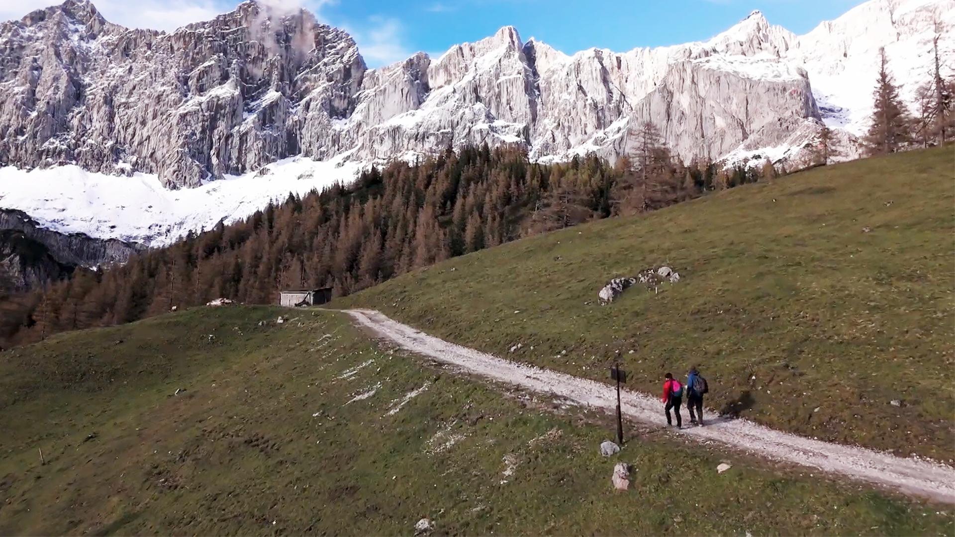 Das Bild zeigt eine alpine Landschaft in Österreich, insbesondere den Dachstein-Gletscher. Im Vordergrund verläuft ein schmaler, erdiger Wanderweg, auf dem zwei Personen mit Rucksäcken gehen. Eine der Personen trägt ein rotes Oberteil, die andere ein blaues.   Links unten ist eine kleine, rustikale Hütte zu sehen, die von Bäumen umgeben ist. Im Hintergrund erhebt sich eine beeindruckende Bergkette mit steilen, zerklüfteten Felsen, die teilweise von Schnee bedeckt sind. Der Himmel ist klar und blau, mit wenigen weißen Wolken. Diese Szenerie vermittelt ein Gefühl von Weite und Naturverbundenheit, passend zu der beschriebenen Reise von den Gletschern zu den Weinbergen in der Steiermark.
