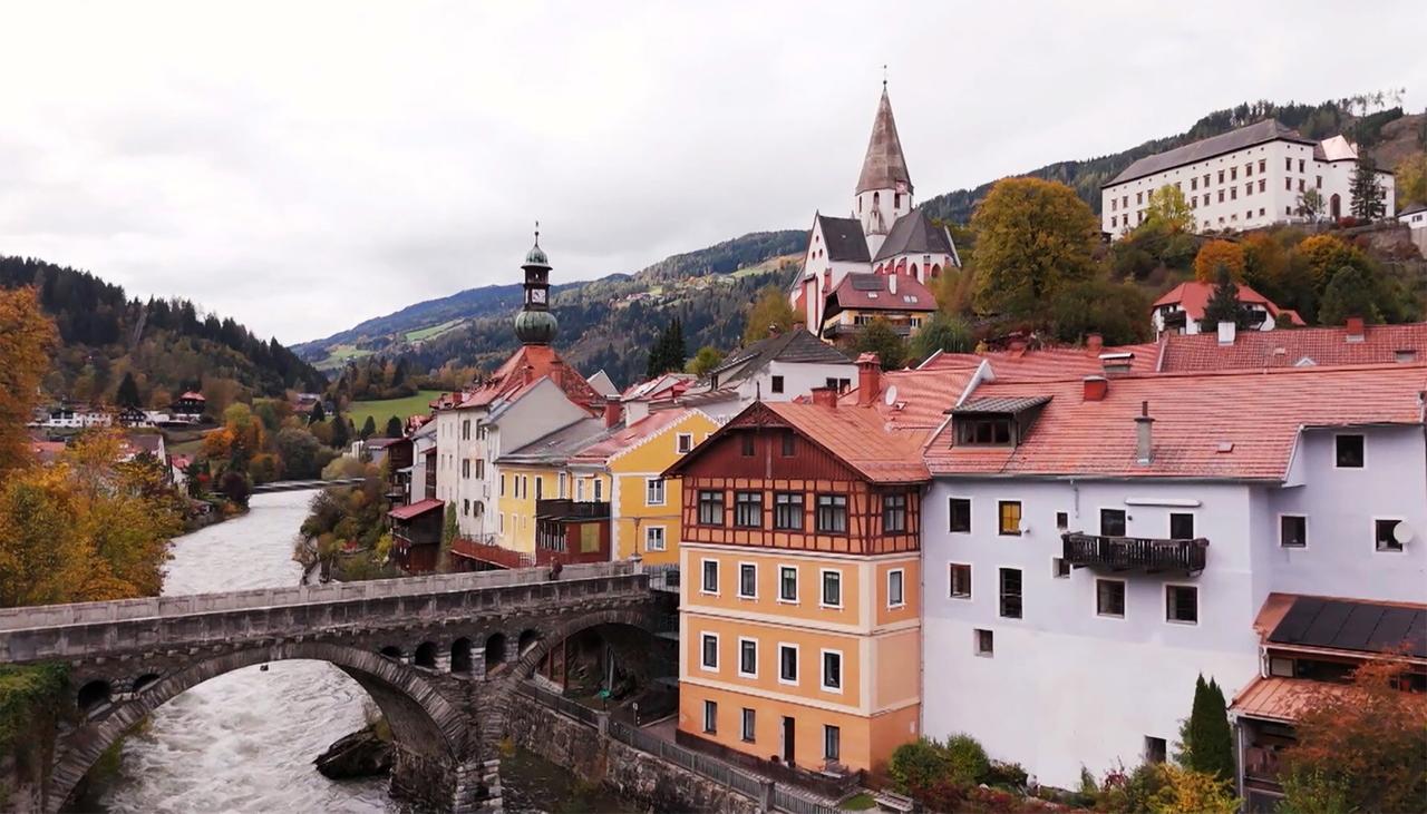 Das Bild zeigt eine pittoreske Stadt im österreichischen Murau. Im Vordergrund verläuft ein Fluss, über den eine steinerne Brücke führt. Die Brücke hat einen gewölbten Bogen und ist aus grauem Stein. Auf der rechten Seite des Bildes sind mehrere Häuser zu sehen, die in verschiedenen Farben gestrichen sind, darunter Gelb, Weiß und Orange. Die Dächer sind mit roten Ziegeln gedeckt.   Im Hintergrund erhebt sich eine Kirche mit einem spitzen Turm und einem charakteristischen Dachtrauf, das mit einer Kuppel endet. Der Landschaftshorizont ist durch sanfte Hügel mit herbstlichen Bäumen geprägt, deren Blätter in warmen Farben leuchten. Der Himmel ist bewölkt, was eine gedämpfte Lichtstimmung erzeugt.   Das gesamte Bild vermittelt eine ruhige, ländliche Atmosphäre und spiegelt die natürliche Schönheit der Region wider.