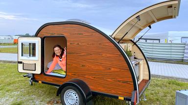 Eine Frau blickt aus der Tür eines Mini-Wohnwagens nach draußen. Im Hintergrund sind abgestellte Wohnwägen zu sehen.