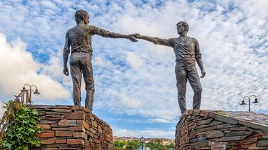 Denkmal zweier Männer, die auf Mauern stehend, sich über den dazwischenliegenden Graben hinweg die Hände reichen.