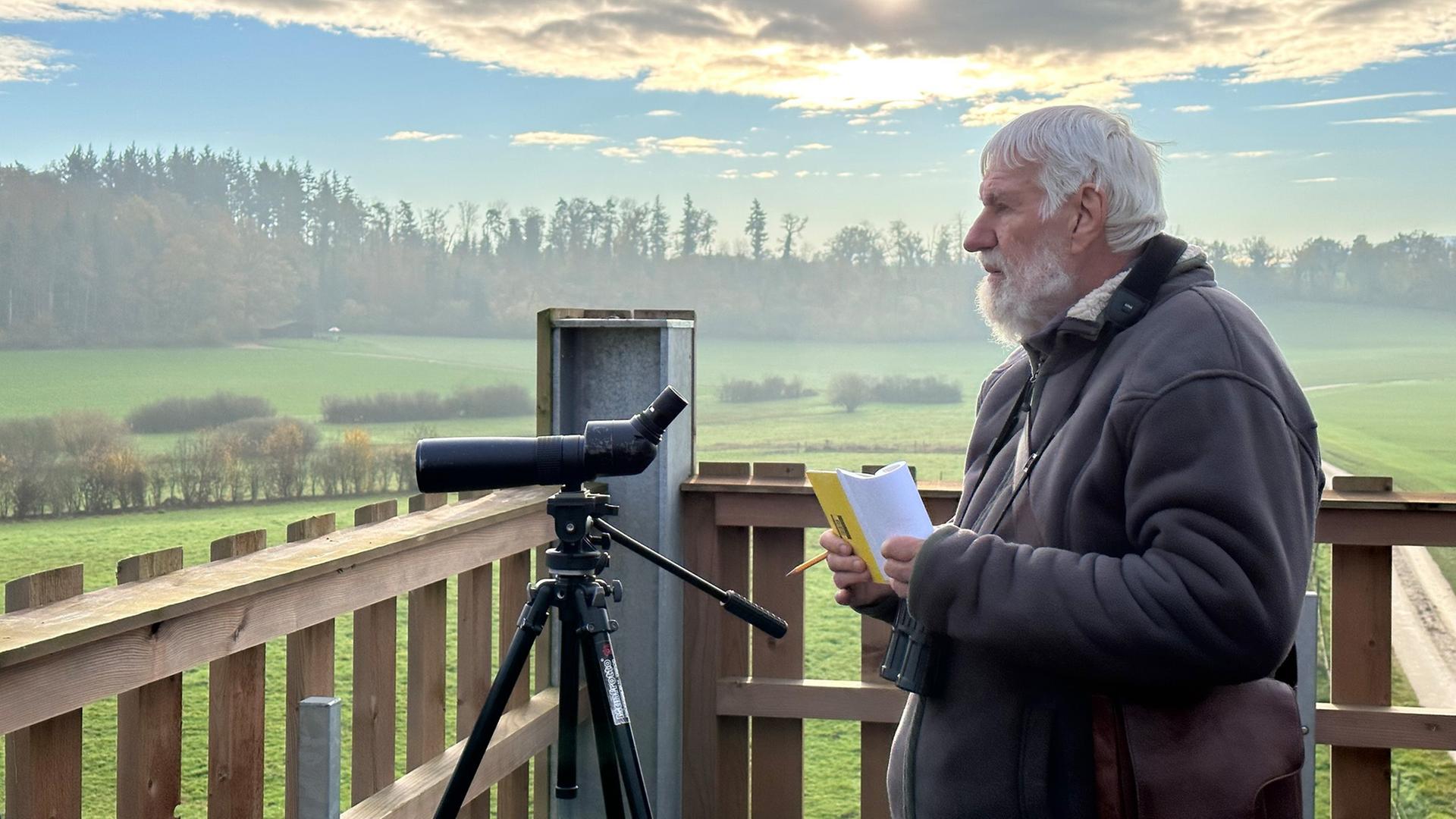 Ein älterer Mann beobachtet mit einem Fernrohr die Landschaft und hält dabei ein Notizbuch in der Hand.