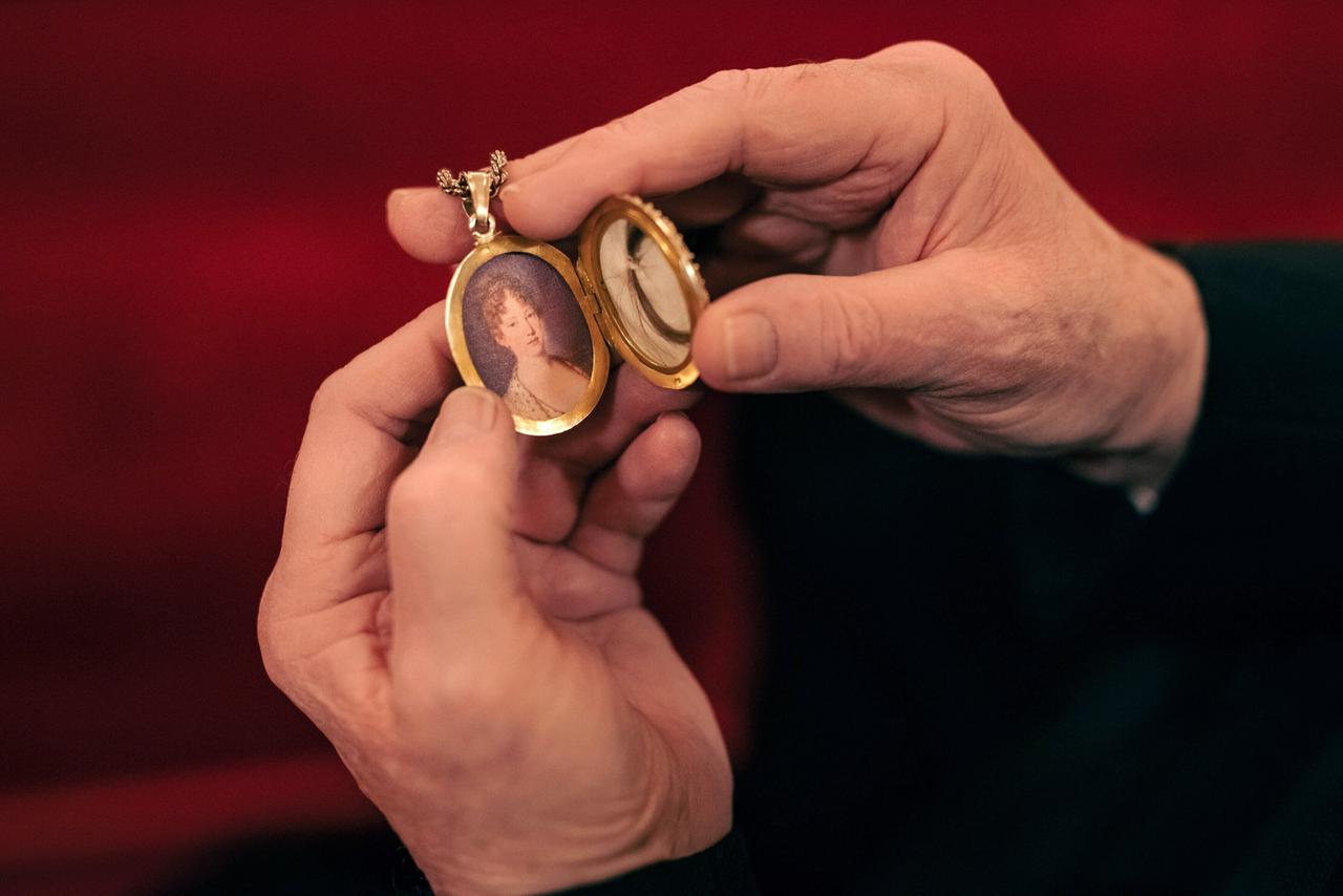 Das Bild zeigt eine Person, die ein Medaillon in den Händen hält. Das Medaillon ist oval und geöffnet, sodass der Blick auf ein Porträt gerichtet ist. Das Porträt zeigt eine historische Figur, die vermutlich Leopoldina Habsburg darstellt, wie aus dem Kontext des Bildes hervorgeht. Der Hintergrund des Bildes ist rot und weich, was eine warme Atmosphäre schafft. Die Hände, die das Medaillon halten, sind gut sichtbar und zeigen Details wie die Falten und Texturen der Haut.