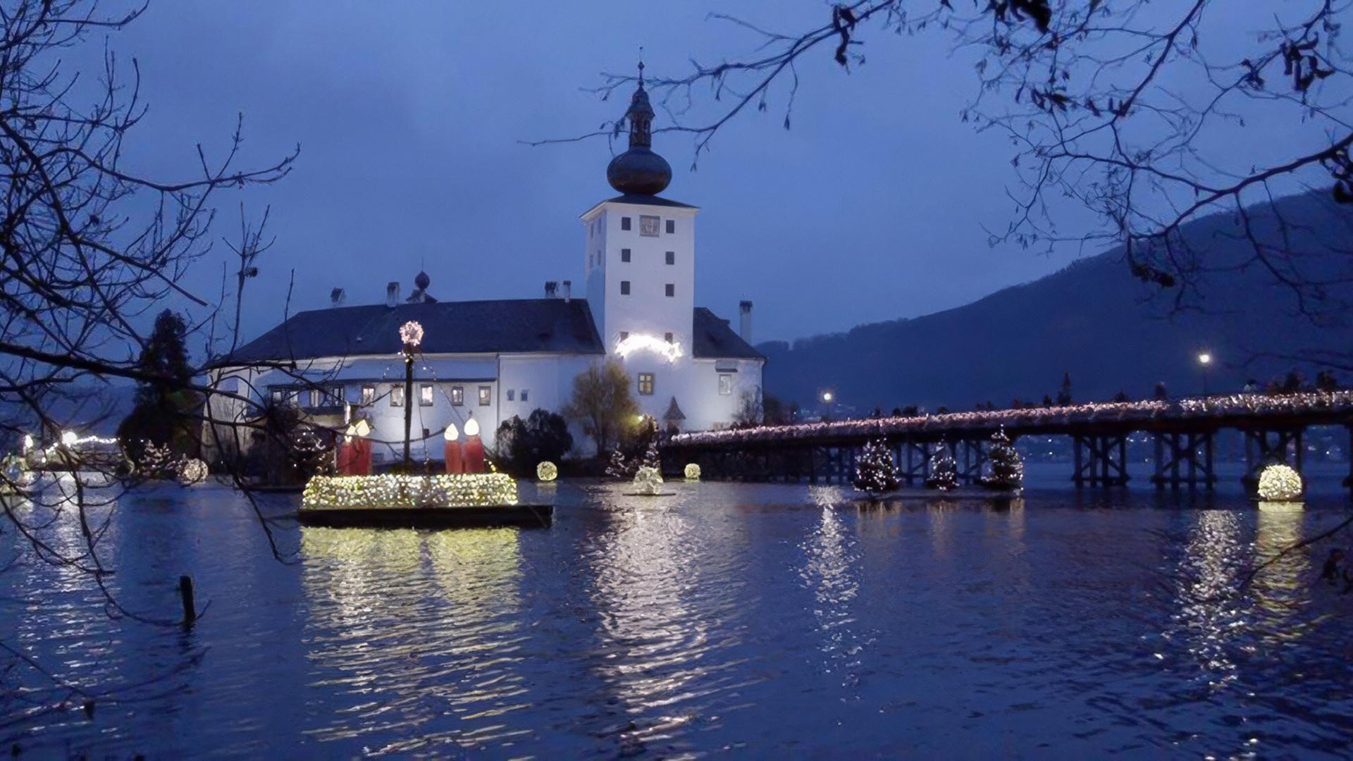 Das Bild zeigt einen stimmungsvollen Abend im Advent, fokussiert auf das Schloss Ort in Gmunden, Österreich. Das Schloss steht auf einer kleinen Insel und ist von Wasser umgeben. Es ist erhellt mit vielen Lichtern, die eine festliche Atmosphäre schaffen.   Im Vordergrund sieht man den dunklen Wasserspiegel, der die Lichter des Schlosses reflektiert. An Land stehen mehrere große, dekorierte Laternen und weihnachtliche Figuren. Ein beleuchteter Steg führt vom Ufer zum Schloss, auf dem Leute flanieren. Der Himmel ist in einem tiefen Blau gehalten und es sind einige Bäume im Vordergrund sichtbar, die die Szenerie umrahmen.  Die Gesamtstimmung ist festlich und besinnlich, typisch für die Adventszeit.