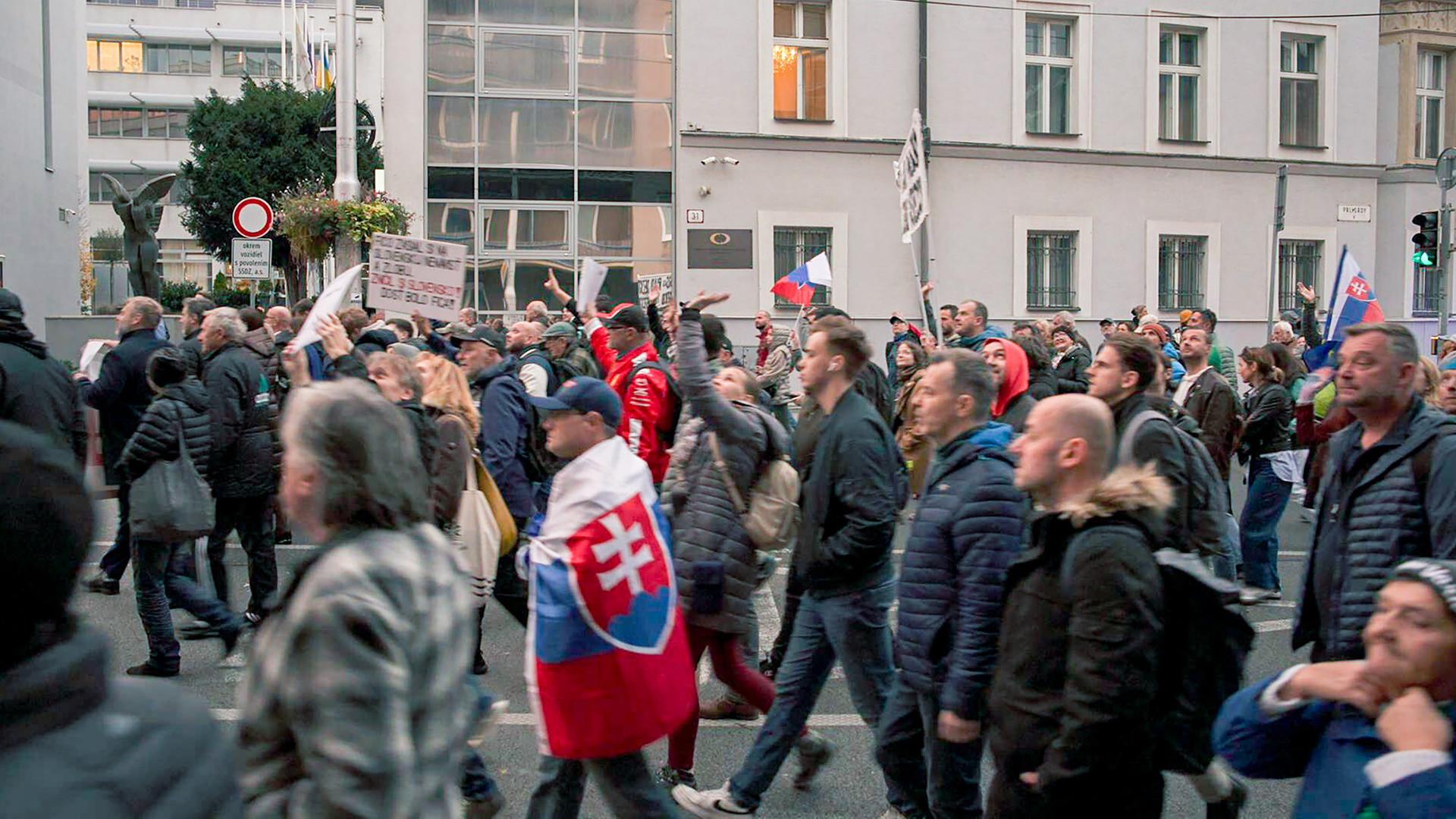 Das Bild zeigt eine Menschenmenge, die auf einer Straße demonstriert. Die Teilnehmerinnen und Teilnehmer tragen überwiegend dunkle winterliche Kleidung und einige von ihnen halten Schilder in den Händen. Es sind auch Fahnen sichtbar, darunter eine mit dem slowakischen Wappen.   Im Hintergrund sind moderne Gebäude zu sehen, darunter Glasfassaden, die das Licht reflektieren. Es wirkt, als wäre die Stimmung in der Menschenmenge aktiv und engagiert, da viele Personen ihre Arme heben und mit Schildern gestikulieren.   Die Szene spielt sich in einer städtischen Umgebung ab, wahrscheinlich in einer Hauptstadt, und deutet auf eine politische Demonstration hin, die sich gegen die Regierung richtet. Die Architektur der Gebäude ist schlicht, mit strukturierten Wänden und einer Mischung aus modernen und traditionellen Elementen.