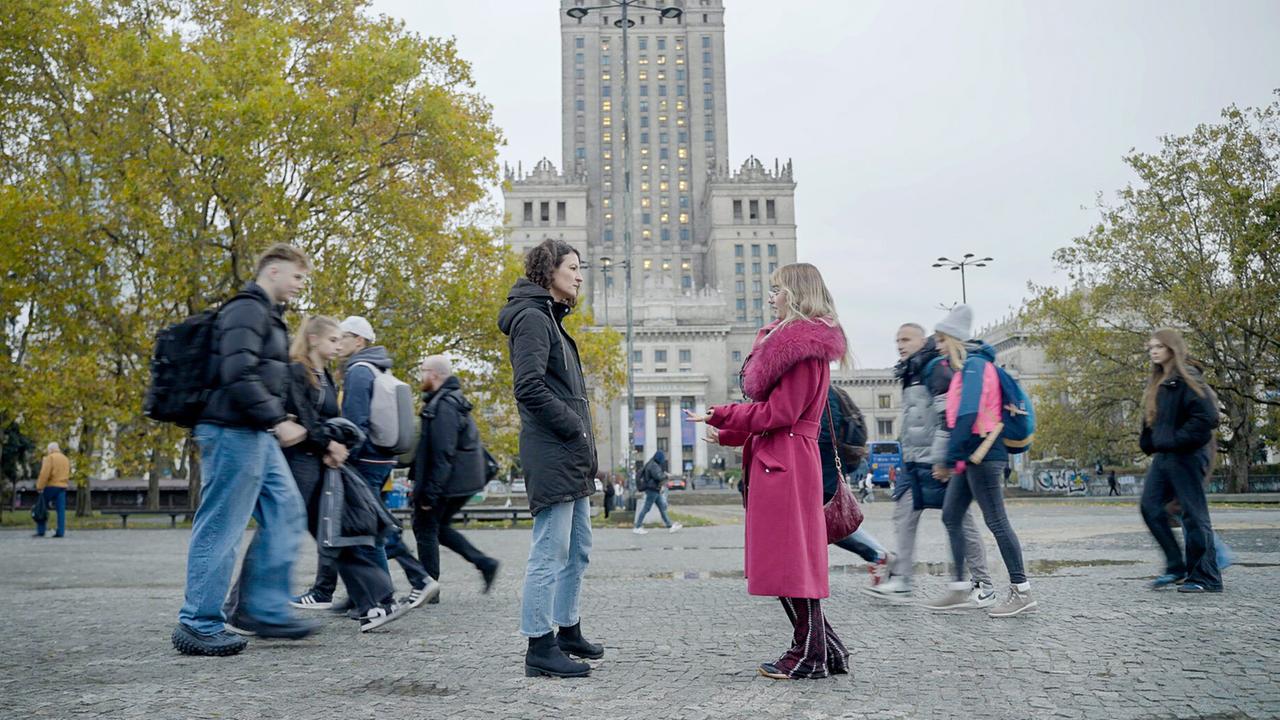 Das Bild zeigt eine belebte Straßenszene in einer urbanen Umgebung, wahrscheinlich in Warschau, Polen. Im Vordergrund stehen zwei Frauen im Gespräch. Die eine trägt einen schwarzen Mantel und hat lockige Haare, während die andere in einen auffälligen, pinken Mantel mit einem rosa Kragen gekleidet ist. Sie scheint gestikuliert und diskutiert ein Thema.  Im Hintergrund ist ein großes, monumentales Gebäude zu sehen, das an die Architektur klassischer sozialistischer Bauweise erinnert. Verschiedene Szenen von Passanten sind sichtbar, darunter eine Gruppe von Jugendlichen und ältere Menschen, die in verschiedene Richtungen gehen, einige tragen Rucksäcke. Die Bäume um die Szene herum haben gelbe Blätter, was auf Herbst hinweist. Der Himmel ist grau und bewölkt, was eine leicht triste Atmosphäre vermittelt.   Die gesamte Komposition vermittelt ein Gefühl von Bewegung und Dynamik in der Stadt, während das Gespräch zwischen den zwei Frauen im Vordergrund eine persönliche Note hinzufügt.