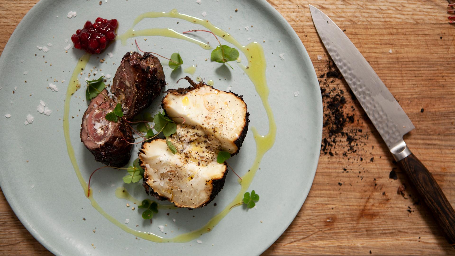 Eine Roulade vom Hirsch, mit Hüttenkäse an Latschenkieferkaramell auf einem Teller. Daneben ein Messer.