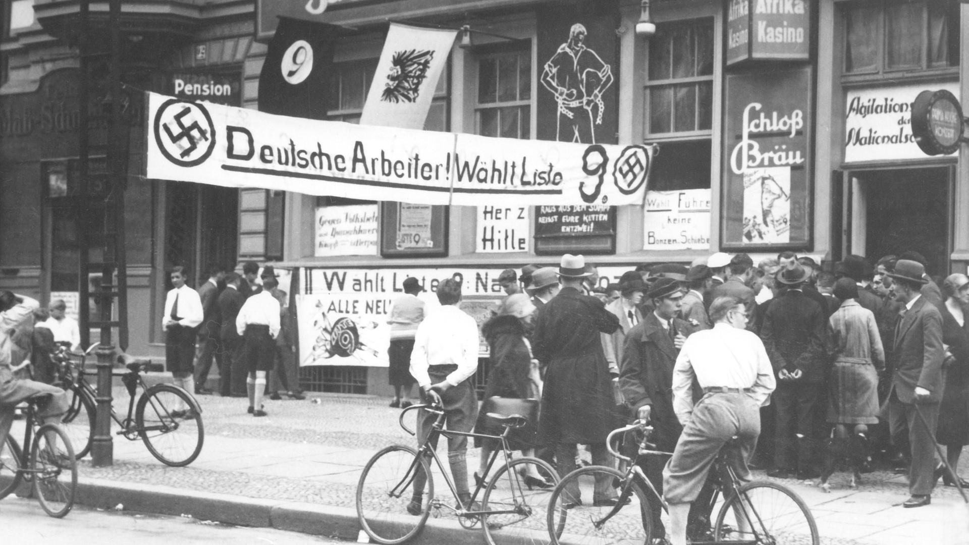 Menschen stehen vor einem Gebäude in Berlin, vor dem ein Banner hängt mit Hakenkreuz und der Wahlwerbung: "Deutsche Arbeiter! Wählt Liste 9"