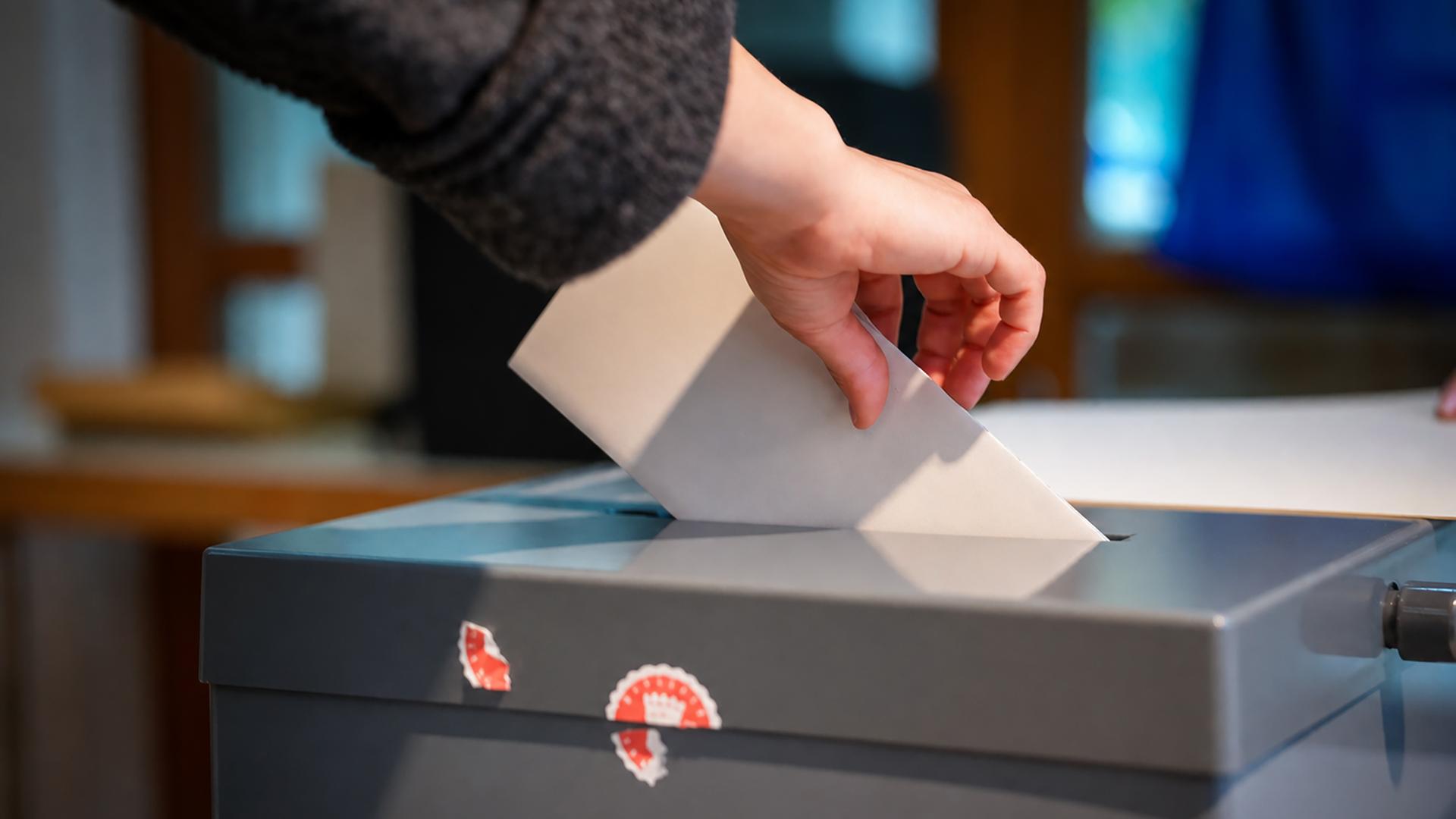 Eine Hand schiebt ein gefaltetes Papier in den Schlitz einer Wahlurne