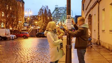 Sarah, eine Frau in einer hellen Jacke und einem Kleinkind auf dem Arm, und Paul, ein Mann in einer dunklen Jacke, kleben ein Suchplakat mit Abreißzetteln an einen Laternenmast. Auf dem Plakat steht "Wohnung dringend gesucht". Neben der Laterne verläuft eine Straße mit Kopfsteinpflaster an der Autos parken. Dahinter steht eine blaue Metallbrücke ("Das Blaue Wunder"). Sarah und das Kind blicken auf das Plakat, während Paul es anbringt.