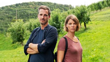 Zwei Kommissare stehen auf einer grünen Wiese mit Apfelbäumen, im Hintergrund bewaldete Hügel  beide blicken ernst in die Kamera.