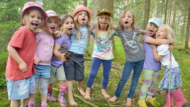 Acht Kinder stehen nebeneinander im Wald, blicken Richtung Kamera und strecken fröhlich die Zunge heraus.