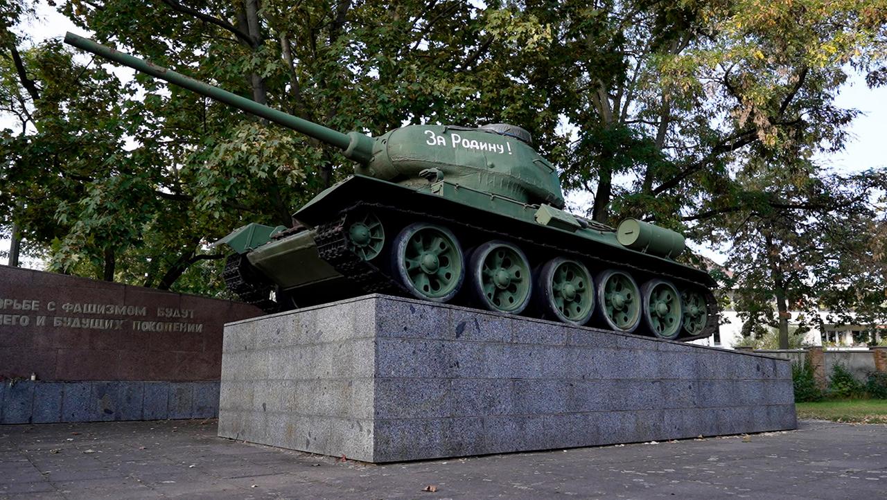 Ein dunkelgrüner Panzer auf einem grauen Steinpodest vor Bäumen