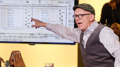 Das Bild zeigt eine Szene aus einer Fernsehshow. Im Vordergrund steht ein runder Tisch mit zwei kleinen Lebkuchenhäusern, die dekoriert sind. Eines ist halbrund und das andere hat eine spitze Dachform. Auf dem Tisch steht zudem ein Glas Wasser und ein Stift.  Hinter dem Tisch ist ein großer Bildschirm zu sehen, auf dem ein Dokument dargestellt ist, das anscheinend Daten oder Namen auflistet.   Ein Mann, der aufgeregt wirkt, steht an dem Tisch und gestikuliert mit einer Hand, während er auf den Bildschirm zeigt. Er trägt ein Hemd mit einem blumigen Muster, eine graue Weste und eine Mütze. Im Hintergrund sind mehrere Zuschauer unscharf zu erkennen, die die Szene beobachten. Die Beleuchtung ist warm und einladend.