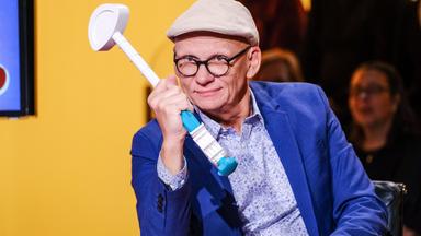 Auf dem Bild ist ein Mann zu sehen, der einen hellen Hut trägt und eine Brille mit schwarzem Gestell hat. Er sitzt an einem Tisch und hält einen Gegenstand in der Hand, der aussieht wie ein Spielzeug oder ein Mikrofon mit einer blauen und weißen Farbe. Der Mann trägt ein blaues Sakko über einem hellen, gemusterten Hemd. Im Hintergrund ist eine wandfarbene gelb-orange Farbe zu erkennen, sowie einige undeutliche Silhouetten von weiteren Personen. Der Gesamtkomplex vermittelt eine fröhliche und lockere Atmosphäre.