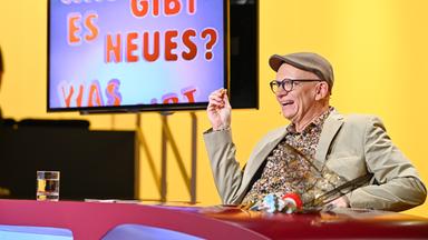 Das Bild zeigt eine Fernsehszene aus der Show "Was gibt es Neues?". Im Vordergrund sitzt ein Mann, der fröhlich aussieht und in einem beigen Anzug mit einem bunten, gemusterten Hemd und einer grauen Mütze gekleidet ist. Er hält in einer Hand einen kleinen Snack und gestikuliert, während er lacht oder spricht.   Hinter ihm ist ein großer Bildschirm zu sehen, auf dem in bunten Buchstaben der Text „WAS GIBT ES NEUES?“ zu lesen ist. Der Hintergrund der Szene ist in warmen Gelbtönen gehalten. Auf dem Tisch vor dem Mann steht ein Glas mit klarem Wasser. Die gesamte Atmosphäre wirkt freundlich und unterhaltsam.