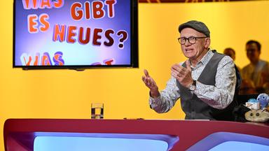 Das Bild zeigt eine Studio-Szene aus der Fernsehsendung "Was gibt es Neues?". Im Vordergrund sitzt ein Mann, der Oliver Baier ist. Er trägt eine graue Mütze, eine brille und ein gemustertes Hemd unter einer grauen Weste. Seine Hände sind erhoben, als ob er einen Punkt erklärt. Vor ihm steht ein Tisch, auf dem ein Glas Wasser und ein Schreibgerät liegen. Rechts auf dem Tisch sind einige farbige Plüschfiguren zu sehen.  Im Hintergrund ist ein Bildschirm, auf dem der Text "Was gibt es Neues?" in bunten Buchstaben zu sehen ist. Die Wand hinter Oliver Baier hat einen gelben Farbton mit einem abstrakten Muster. Im Hintergrund sind einige schemenhafte Personen zu erkennen, die im Publikum sitzen.   Die Gesamtatmosphäre vermittelt eine lebhafte und unterhaltsame Stimmung, die typisch für eine Quiz- oder Unterhaltungssendung ist.