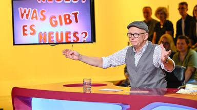 Das Bild zeigt eine Fernsehsendung mit dem Titel „Was gibt es Neues?“. Im Vordergrund steht ein Moderator, der Oliver Baier ist. Er trägt eine graue Weste über einem weißen Hemd mit buntem Muster und eine graue Schirmmütze. Seine Brille hat dünne, schwarze Rahmen. Er gestikuliert mit einer Hand, während er auf einem Tisch sitzt, der eine rote Oberfläche hat. Auf dem Tisch steht ein Glas mit Wasser und einige Unterlagen.  Im Hintergrund ist ein Bildschirm sichtbar, der den Titel der Show in bunten Buchstaben anzeigt: „NEUES? WAS GIBT ES NEUES?“. Der Hintergrund des Bildes ist gelb. Hinter dem Moderator sind mehrere Zuschauer zu sehen, die teilweise unscharf sind. Sie scheinen interessiert zuzuschauen.