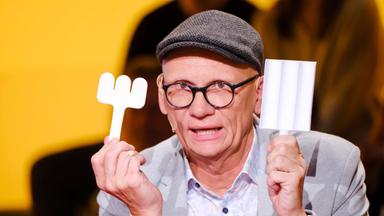 Oliver Baier mit Brille und Schirmmütze hält zwei skurrile weiße Gegenstände in die Kamera, im Hintergrund ein gelber Studiowand und unscharfe Zuschauer.