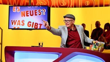Ein Mann mit grauem Blazer und einer Kappe in einer gelb beleuchteten Bühnendeko an einem Tisch sitzend.