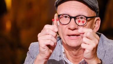 Oliver Baier trägt eine Schirmmütze und Brille und hält zwei rote Quiz-Objekte mit Drahtgestell vor sein Gesicht; er spricht in einem Studio mit warmer Beleuchtung.