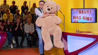 Auf dem Bild ist ein Moderator zu sehen, der einen großen, plüschigen Teddy in den Armen hält. Der Moderator trägt ein Hemd mit einem bunten Muster, eine Weste und eine Mütze. Im Hintergrund sitzt ein Publikum auf gestuften Plätzen und applaudiert.  Das Setting ist eine Fernsehsendung mit einem farbenfrohen, modern gestalteten Set. An einer Wand hinter dem Moderator ist ein Bildschirm mit dem Schriftzug „ES NEUES? WAS GIBT ES“ zu sehen. Der Tisch, an dem der Moderator steht, hat eine kurvenreiche, rote Oberfläche und ist teilweise transparent blau beleuchtet. Auf dem Tisch liegen einige Notizen und ein Glas.   Die gesamte Szenerie strahlt eine fröhliche und unterhaltsame Atmosphäre aus.