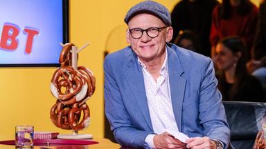 Das Bild zeigt einen Mann, der am Tisch vor einem orangefarbenen Hintergrund sitzt. Er trägt eine graue Mütze und eine Brille mit dicken, schwarzen Rahmen. Seine Kleidung besteht aus einem hellblauen Blazer über einem weißen Hemd mit bunten Punkten. Auf dem Tisch vor ihm liegen einige Zettel und ein Glas mit Wasser.  Neben ihm steht eine Art Ständer mit mehreren Brezeln, die übereinander gestapelt sind. Im Hintergrund sind mehrere Personen zu sehen, die an einer Art Publikumssitzplatz sitzen. Der Gesamtaufbau der Szene scheint zu einem Fernsehformat zu gehören, das sich mit dem Thema „Brezel“ beschäftigt.