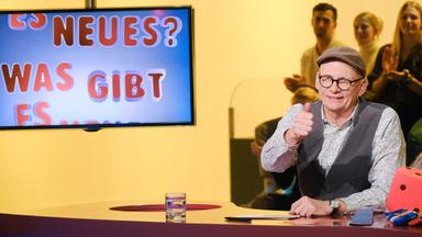 Das Bild zeigt eine Fernsehszene aus der Sendung "Was gibt es Neues?". Im Vordergrund sitzt ein Mann, der Oliver Baier heißt. Er trägt eine graue Kappe, eine Brille mit dunklem Rahmen und hat ein helles, gemustertes Hemd an, über dem er eine dunkle Weste trägt. Er lächelt und gibt mit der rechten Hand ein Daumenhoch. Vor ihm befindet sich ein runder Tisch, auf dem ein Glas mit Wasser steht.   Im Hintergrund ist ein Bildschirm zu sehen, auf dem der Text "ES NEUES? WAS GIBT ES NEUES?" in bunten Buchstaben dargestellt ist. Die Farbgebung im Hintergrund ist überwiegend gelb, und in der Nähe des Tisches sind mehrere Personen zu erkennen, die im Publikum sitzen und applaudieren.