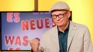 Das Bild zeigt einen Mann, der an einem Tisch sitzt. Er trägt eine graue Jacke, ein weißes Hemd mit einem blau-weißen Muster und eine graue Schirmmütze. Vor ihm steht ein Glas Wasser und er hält einen Stift in der Hand. Im Hintergrund ist ein großer Bildschirm zu sehen, auf dem ein Tier, vermutlich ein Ameisenigel, abgebildet ist. Der Hintergrund des Raumes ist gelb, was eine warmes Lichtambiente schafft. Die Szene scheint aus einer Fernsehsendung zu stammen.