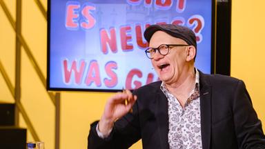Das Bild zeigt eine Szene aus einer Fernsehsendung mit einem Moderator, der in einem Studio sitzt. Der Moderator hat eine Glatze, trägt eine Brille und eine graue Mütze. Er ist lachend abgebildet und hält ein rotes Blatt Papier in der Hand. Hinter ihm ist ein Bildschirm zu sehen, auf dem in großen, bunten Buchstaben steht: "ES GIBT NEUES? WAS GIBT?" Die Hintergrundfarbe des Bildschirms ist blau und gelb. Der Tisch, an dem der Moderator sitzt, hat eine Holzoberfläche mit runden roten Einlagen. Auf dem Tisch steht ein Wasserglas. Die Szene vermittelt eine fröhliche und unterhaltsame Atmosphäre.
