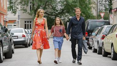 Eine Frau im roten Blumenkleid, ein Mädchen in Shorts und Ringelshirt sowie ein Mann in dunklem Hemd gehen Hand in Hand auf einer ruhigen Stadtstraße spazieren.