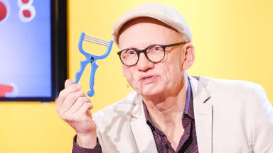 Das Bild zeigt einen Mann, der einen blauen Gemüseschäler in der Hand hält. Er trägt eine helle Jacke und eine Kappe. Der Mann hat eine Brille auf und steht vor einem Hintergrund in Gelb, der mit grafischen Elementen gestaltet ist. Unmittelbar hinter ihm ist ein Bildschirm sichtbar, auf dem teilweise farbige Fragezeichen zu sehen sind. Der Ausdruck des Mannes wirkt konzentriert und er spricht offensichtlich in eine Kamera.