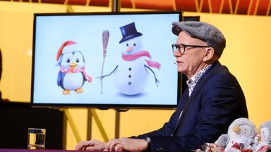 Das Bild zeigt einen männlichen Moderator mit einer schräg sitzenden Mütze und einer Brille. Er sitzt an einem Tisch, auf dem ein Glas Wasser steht. Im Hintergrund befindet sich ein Bildschirm, auf dem zwei cartoonhafte Figuren abgebildet sind: ein Pinguin mit einem roten Schal und einer Weihnachtsmütze sowie ein Schneemann mit einem schwarzen Zylinder und einem roten Schal. Der Tisch ist mit kleinen Dekorationsfiguren von Pinguinen und Schneemännern geschmückt. Der Hintergrund ist in warmen Gelbtönen gehalten, und das Bild vermittelt eine festliche Atmosphäre.