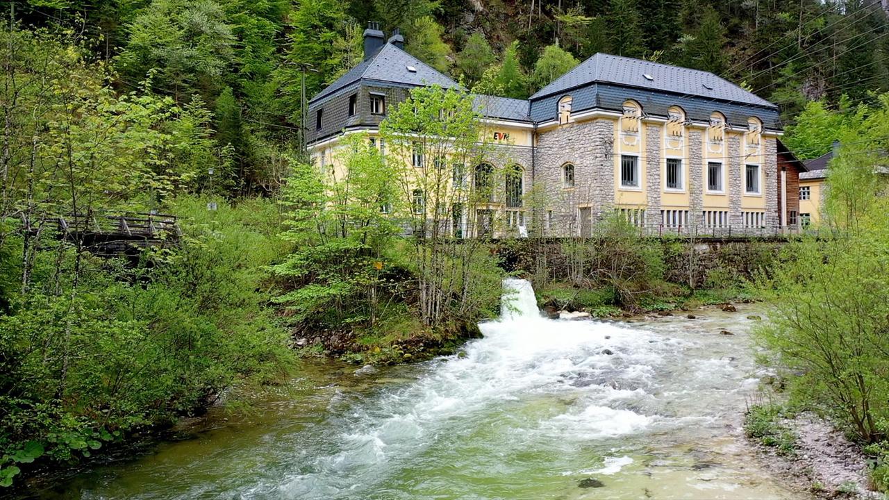 Ein historisches Wasserkraftwerk mit gelbem Mauerwerk liegt eingebettet in einem grünen Tal, davor fließt ein klarer Gebirgsfluss.