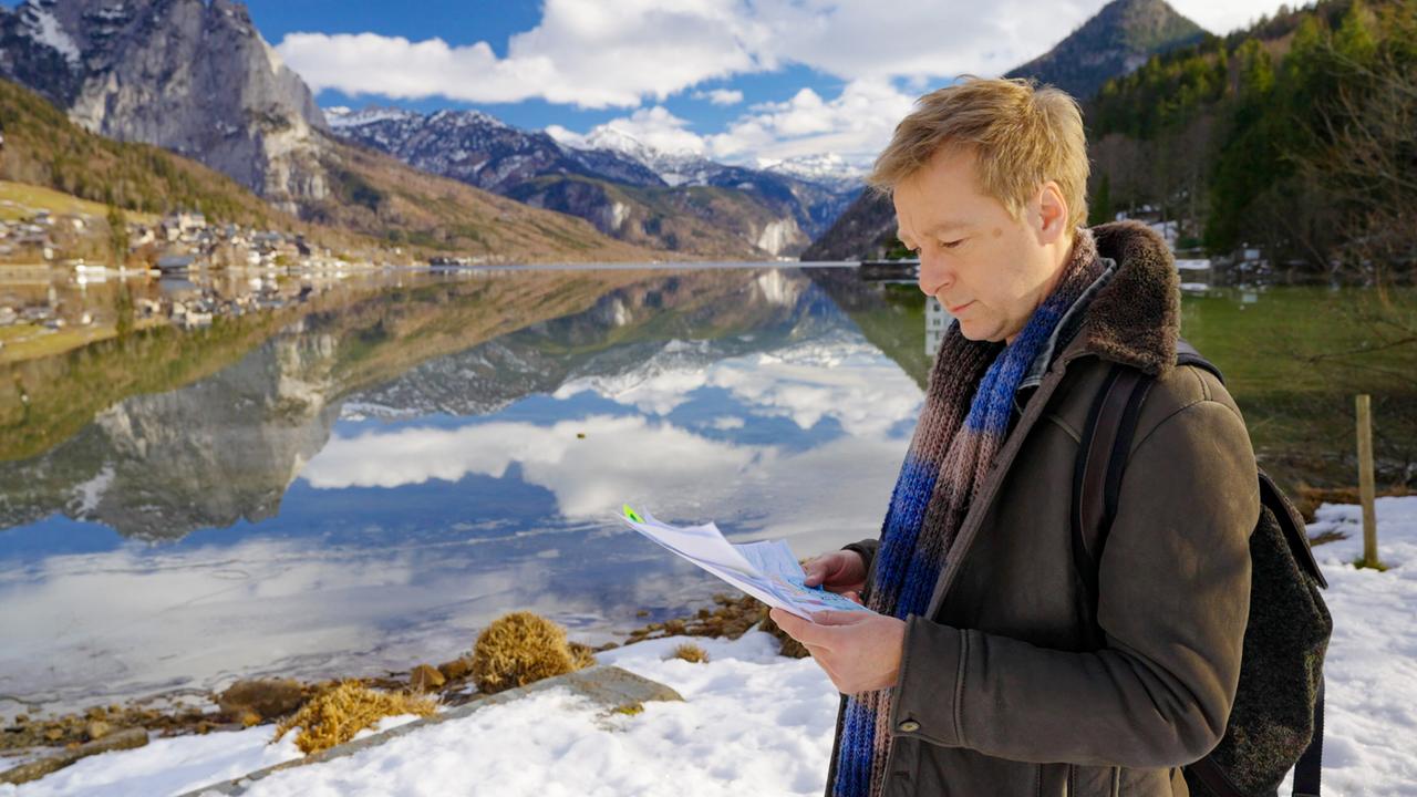Das Bild zeigt eine winterliche Landschaft am Grundlsee, umgeben von hohen Bergen, die teilweise mit Schnee bedeckt sind. Der Himmel ist blau mit einigen weißen Wolken. Im Vordergrund steht ein Mann, vermutlich Max Müller, der einige Dokumente in den Händen hält; er scheint nachdenklich zu sein. Er trägt einen dunklen, gefütterten Mantel und einen bunten Schal. Auf dem Boden befindet sich eine schneebedeckte Fläche, und im Hintergrund spiegelt sich die Landschaft im ruhigen Wasser des Sees wider. Am Ufer sind einige Gräser und/or Pflanzen sichtbar, die aus dem Schnee ragen. Die Atmosphäre ist ruhig und idyllisch, und die Szenerie vermittelt ein Gefühl von Entdeckung und Erkundung.