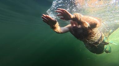 Das Bild zeigt einen Mann, der unter Wasser schwimmt. Er trägt keine Kleidung und hat eine lange, dunkle Haarpracht, die um seinen Kopf und Schultern schwingt. An seinen Handgelenken sind faserige Armbänder zu erkennen. Der Mann streckt seine Arme nach vorne, während er im Wasser vorwärts schwimmt. Die Wasseroberfläche ist darüber sichtbar, wobei Blasen aufsteigen. Das Licht fällt schimmernd auf das Wasser, was eine grüne und verführerische Unterwasserlandschaft schafft. Der Hintergrund ist dunkler, während die Lichtreflexionen an der Oberfläche eine lebendige Atmosphäre erzeugen.