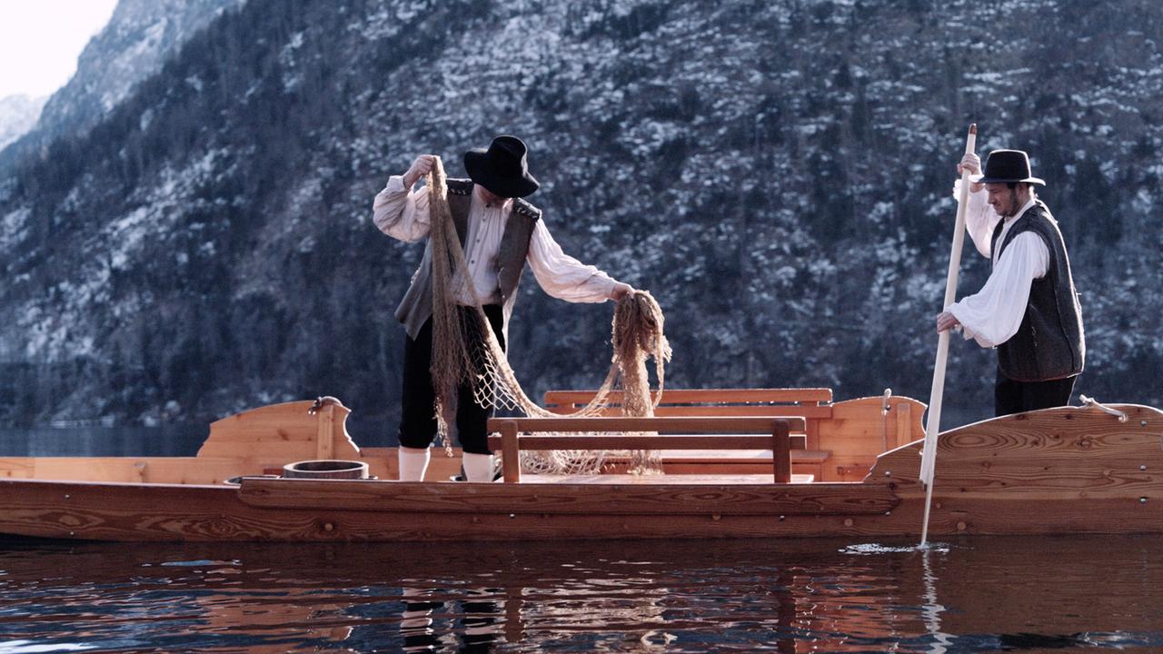 Das Bild zeigt eine Szene, die auf einem See spielt, wahrscheinlich am Grundlsee in Österreich. Zwei Männer in historischen Trachten sind in einem Holzboot.   Der Mann auf der linken Seite hält ein Netz in der Hand, während er es sortiert. Er trägt eine weiße, langärmelige Bluse, eine Weste und einen breiten, schwarzen Hut. Der andere Mann, auf der rechten Seite, steht mit einem langen Stock, der in das Wasser reicht, und hat ebenfalls eine ähnliche Tracht an.   Im Hintergrund sind hohe, schneebedeckte Berge zu sehen, die in den See reflektiert werden. Die Wasseroberfläche ist ruhig, und die gesamte Szene vermittelt eine winterliche Atmosphäre.   Das Bild ist Teil einer Geschichte, die sich mit dem Wassermann vom Grundlsee beschäftigt und die Folklore der Region behandelt.