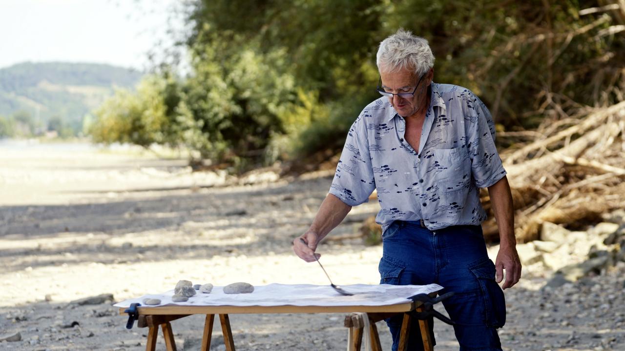 Ein Mann steht an einem Uferbereich und arbeitet an einem Tisch mit einem weißen Tuch. Vor ihm liegen Steine. Im Hintergrund sind Bäume und eine Wasserfläche sichtbar.