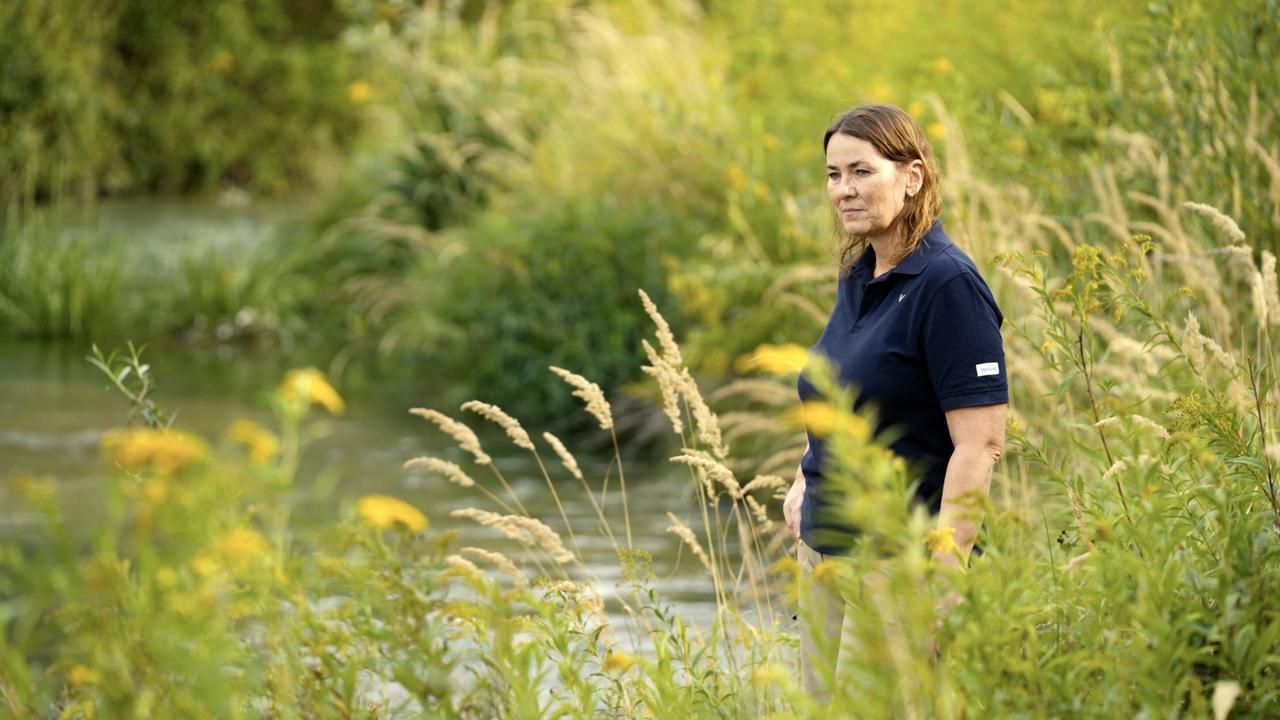 Eine Frau steht am Ufer eines Gewässers, umgeben von hohen Gräsern und wild wachsenden Pflanzen. Im Hintergrund sind Bäume und ein bewaldetes Terrain sichtbar, das die Szenerie umrahmt.