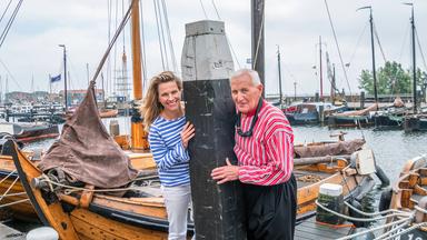 Frau und ein älterer Mann stehen in einem Hafen, zwischen ihnen ein Poller