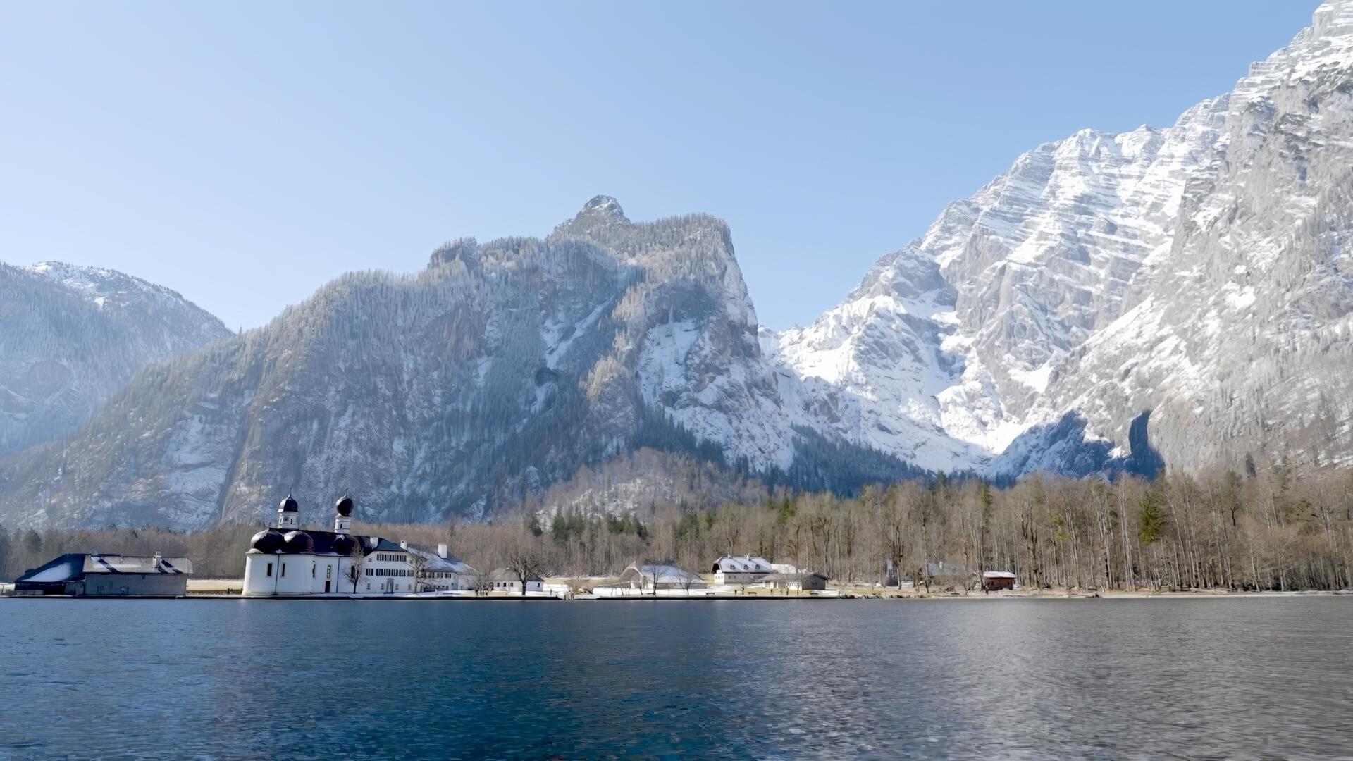 Das Bild zeigt eine winterliche Landschaft im Berchtesgadener Land, mit dem Königssee im Vordergrund und der markanten Ostwand des Watzmann im Hintergrund. Auf der linken Seite des Bildes ist die weithin bekannte Kirche St. Bartholomä zu sehen, eine auffällige, weiße Baufläche mit zwei charakteristischen, runden Türmen. Der See ist ruhig und spiegelnd, was die umliegenden verschneiten Berge reflektiert.  Im Hintergrund erkennt man große, steile Bergformationen mit schneebedeckten Gipfeln, die den Eindruck von majestätischer Erhabenheit erwecken. Die Bäume am Ufer sind kahl, während die Region von Schnee bedeckt ist, was eine ruhige und friedliche Atmosphäre schafft. Der Himmel ist klar und blau, was typisch für kalte Wintertage in den Alpen ist. Das Gesamtbild vermittelt ein Gefühl von natürlicher Schönheit und Klarheit in einer beeindruckenden Berglandschaft.