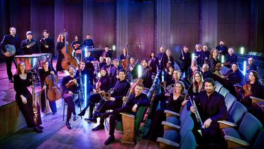 Die Mitglieder des WDR-Funkhausorchesters sitzen lächelnd im Zuschauerraum eines Konzertsaals und halten Musikinstrumente in den Händen.