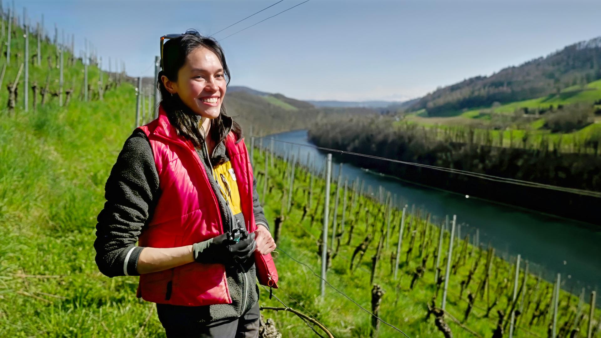 Junge Frau mit roter Weste steht auf einem sonnigen Weinberg oberhalb eines Flusses, im Hintergrund grüne Hügel und Rebenreihen.