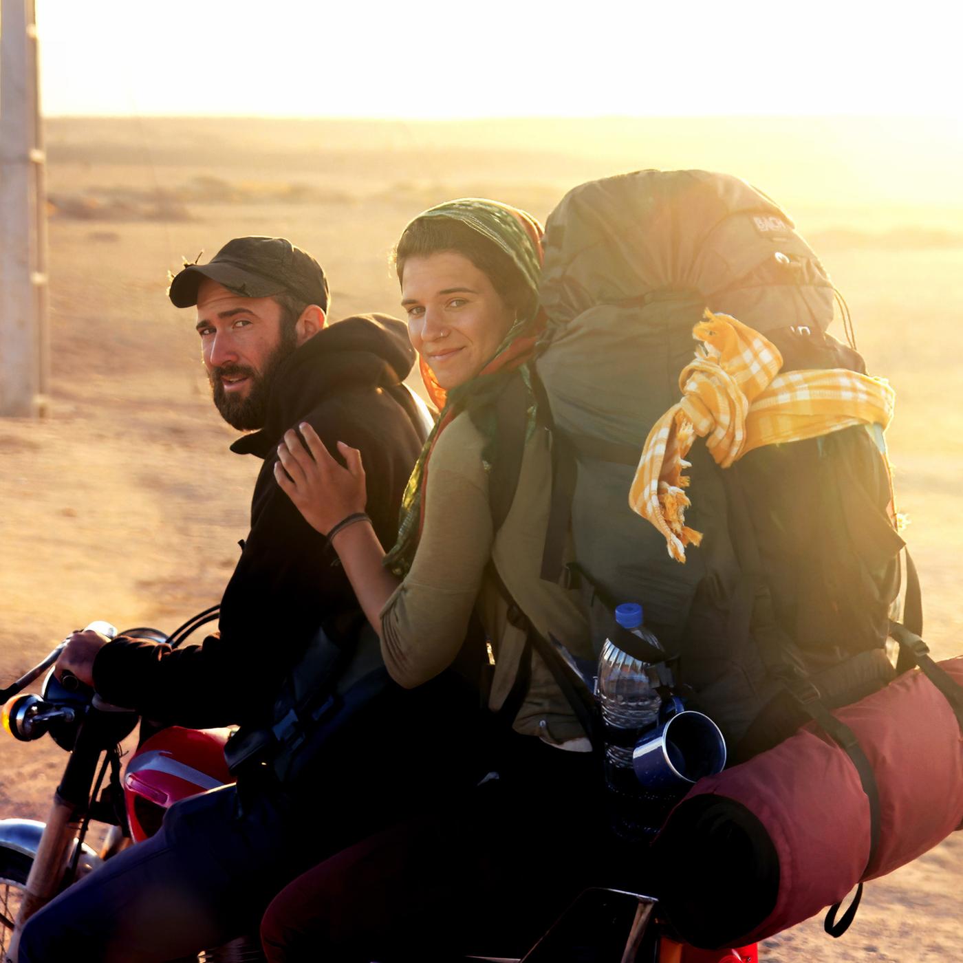 Ein junger Mann mit Bart und Baseballkappe sitzt am Lenker eines Motorrads und schaut in die Kamera. Hinter ihm sitzt eine junge Frau, die sich an seiner Schulter festhält, die Haare mit einem Tuch bedeckt hat und einen vollbepackten Rucksack auf den Schultern trägt. Im Hintergrund ist eine weite Steppenlandschaft unscharf und in einem goldgelben Licht der Abendsonne zu sehen.