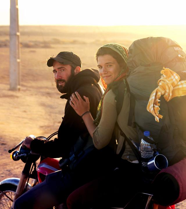 Ein junger Mann mit Bart und Baseballkappe sitzt am Lenker eines Motorrads und schaut in die Kamera. Hinter ihm sitzt eine junge Frau, die sich an seiner Schulter festhält, die Haare mit einem Tuch bedeckt hat und einen vollbepackten Rucksack auf den Schultern trägt. Im Hintergrund ist eine weite Steppenlandschaft unscharf und in einem goldgelben Licht der Abendsonne zu sehen.